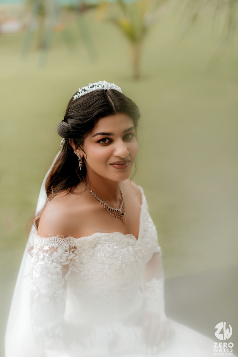 Bride wearing an off-shoulder lace wedding dress with tiara and jewelry in a soft outdoor setting.