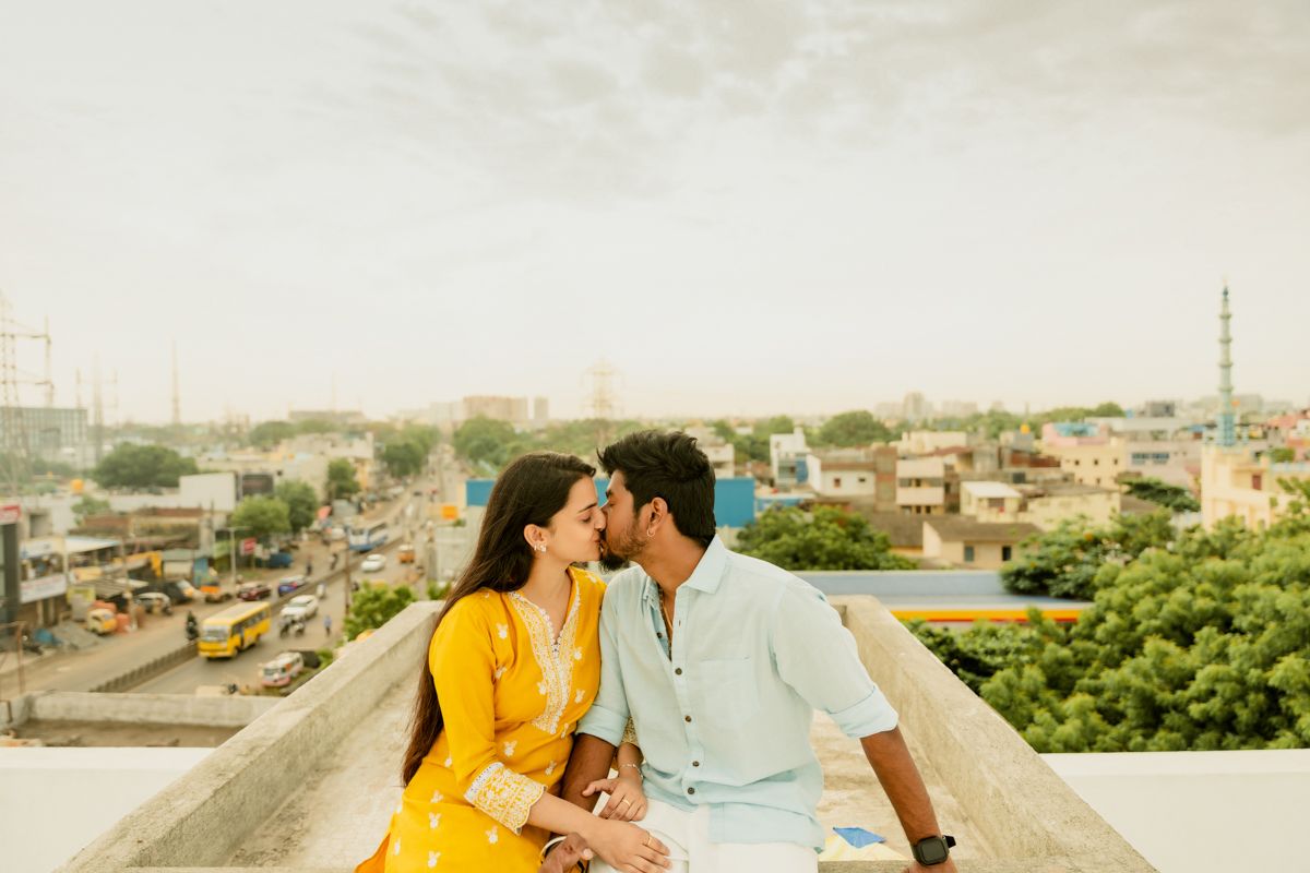 A couple in contrasting yellow and light blue attire embrace while sitting on a rooftop with an urban view behind them.