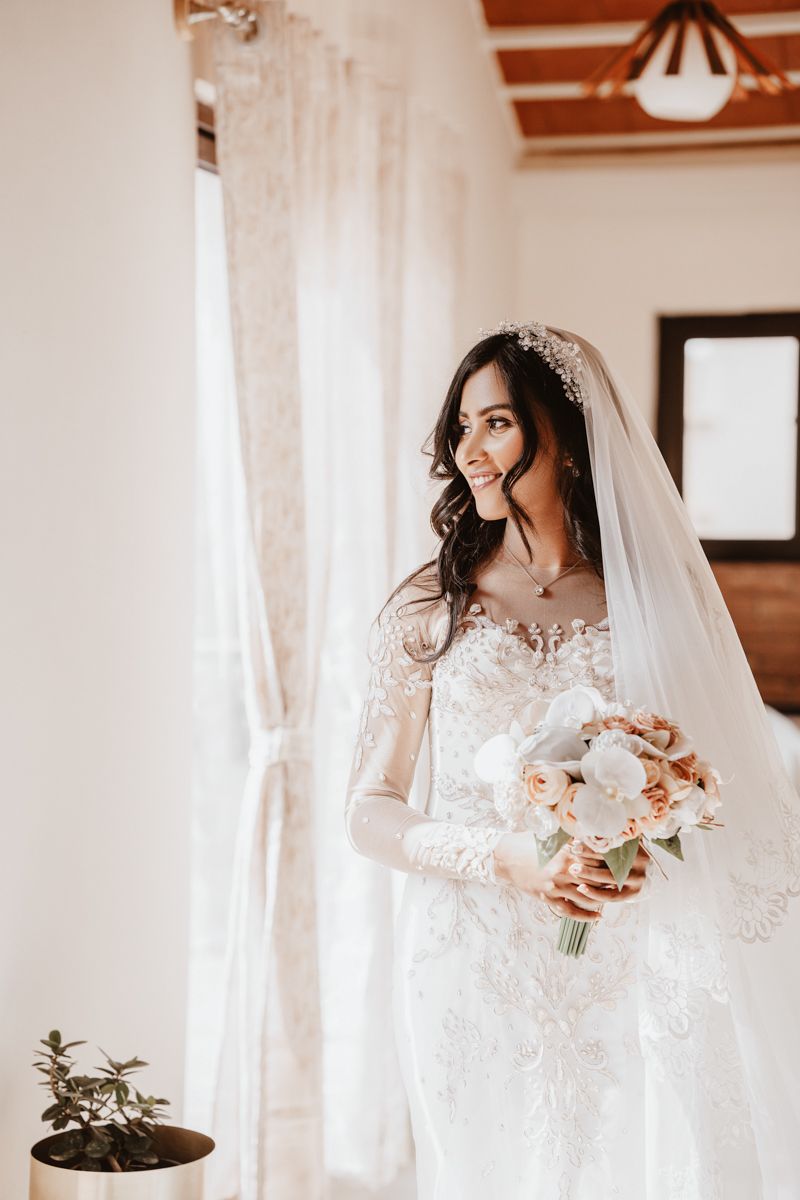 Person in elegant lace wedding dress holding peach and white floral bouquet near sunlit window.