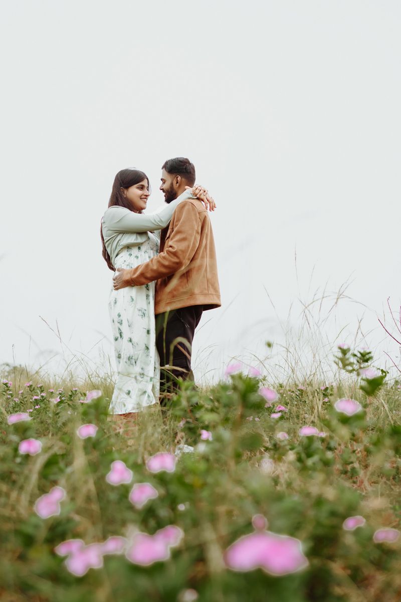 A romantic couple embraces on a hilltop surrounded by delicate pink wildflowers on an overcast day.