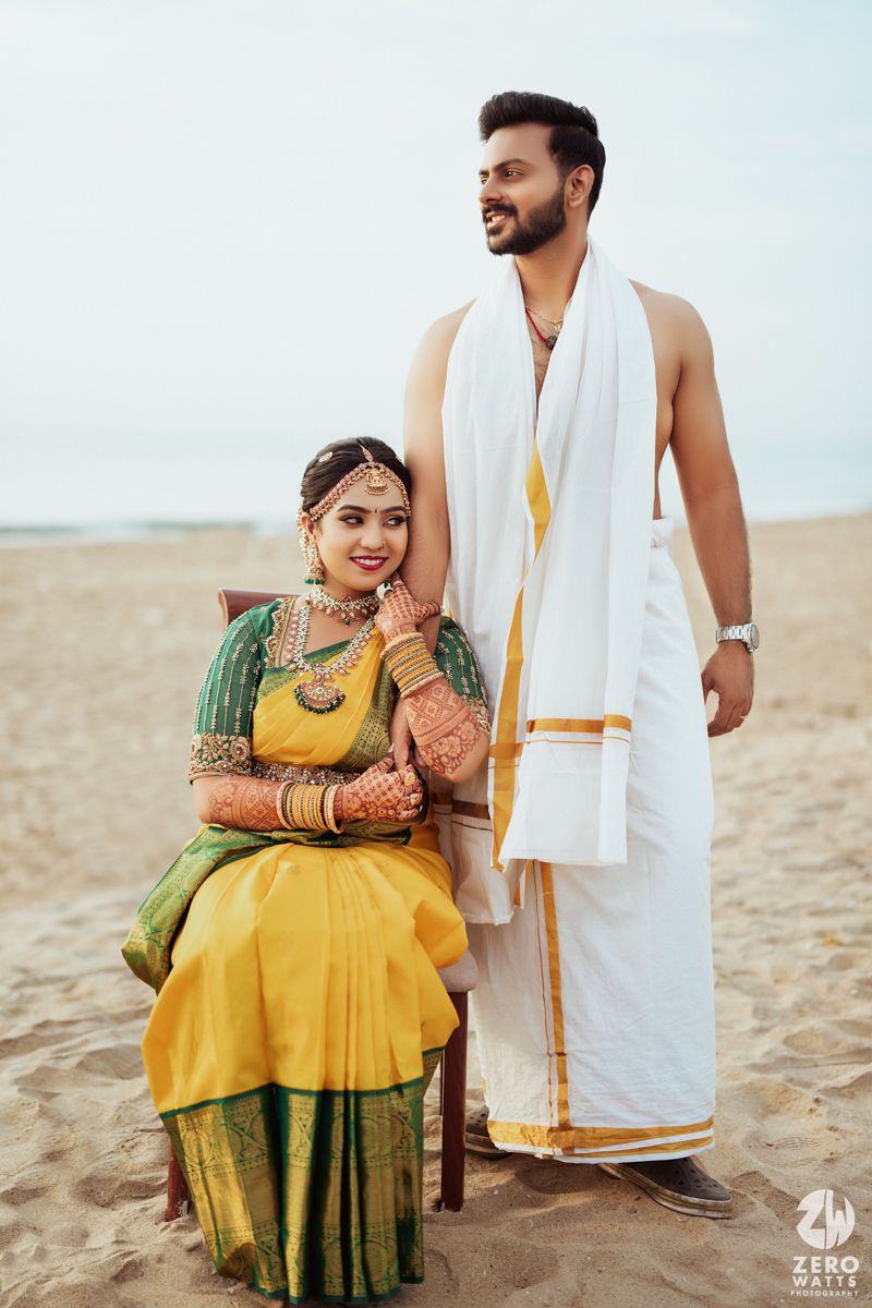 A couple in traditional South Indian wedding attire poses on a beach, with the bride in yellow and green saree.
