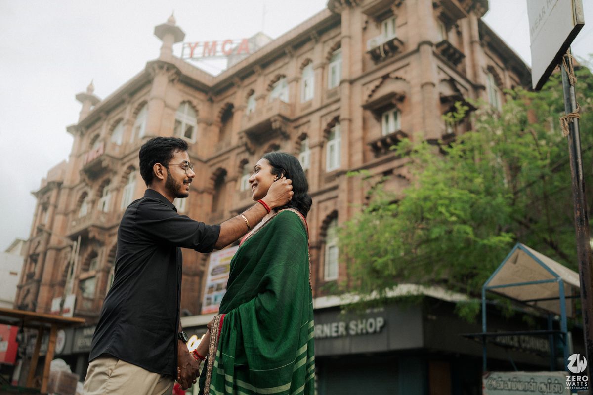 Two people interacting on city street with historic Victorian architecture in background.
