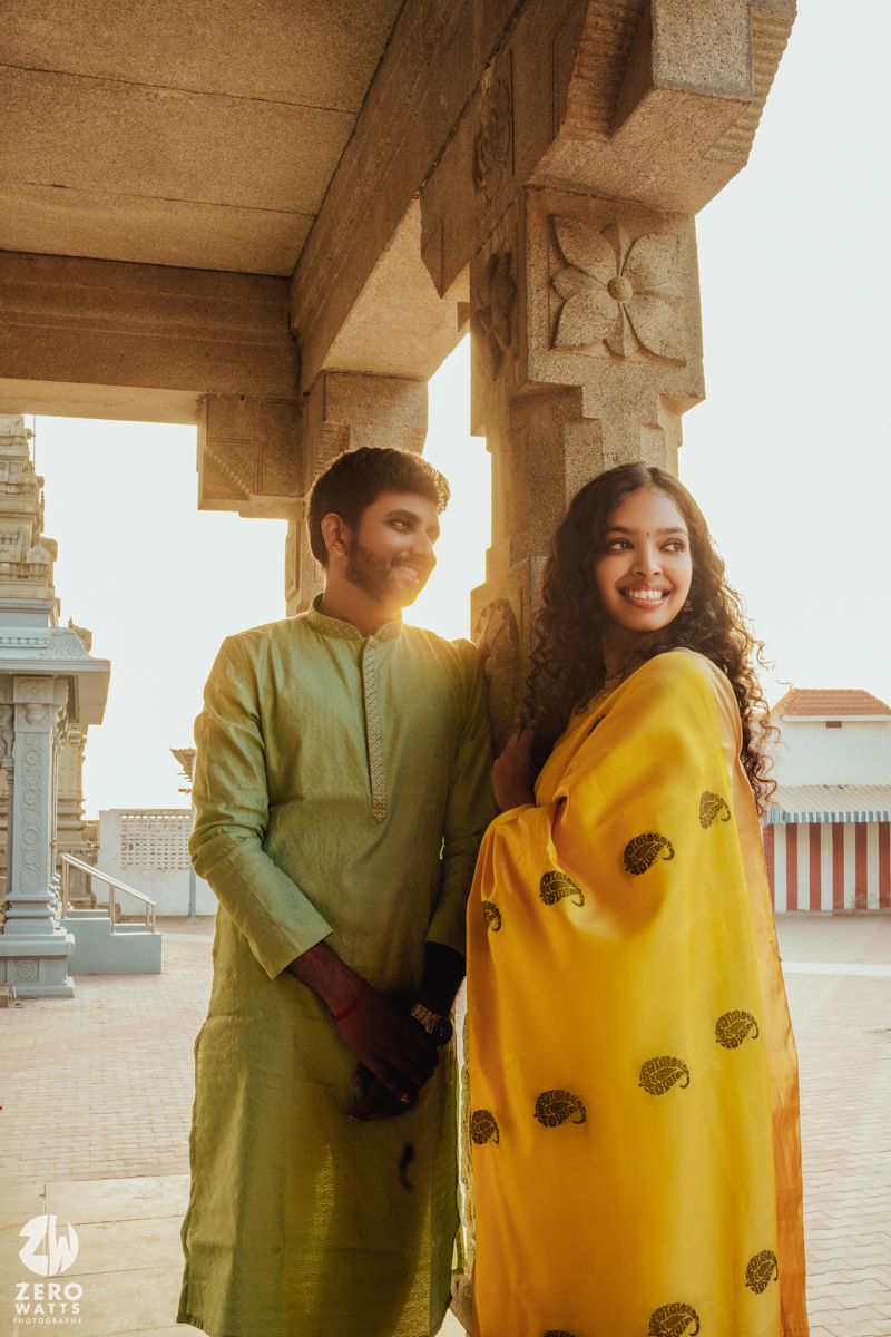 A couple dressed in traditional Indian attire, with yellow sari and olive kurta, pose in a temple corridor at sunset.