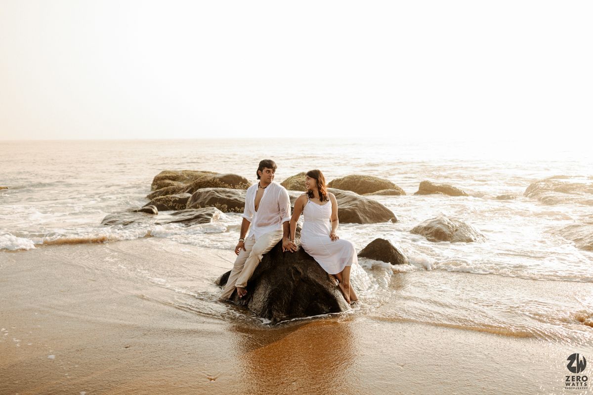 Couple sits together on rocky beach formation during sunset with ocean waves in background.