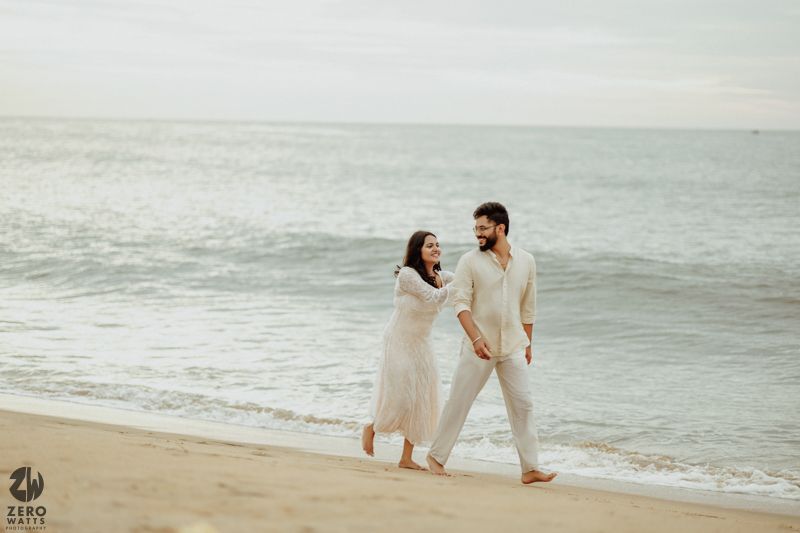 A couple in white outfits walking together along a misty beach shoreline at sunset.