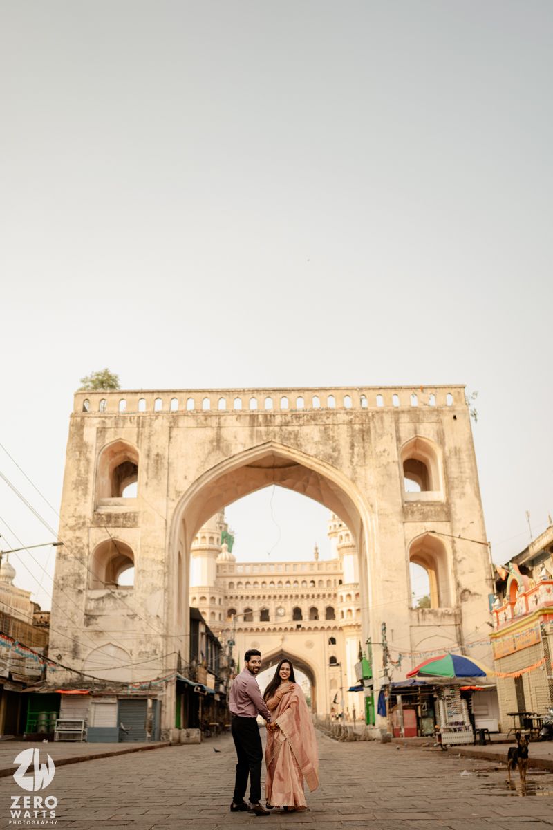 A couple poses in front of a grand archway entrance to a historic monument.