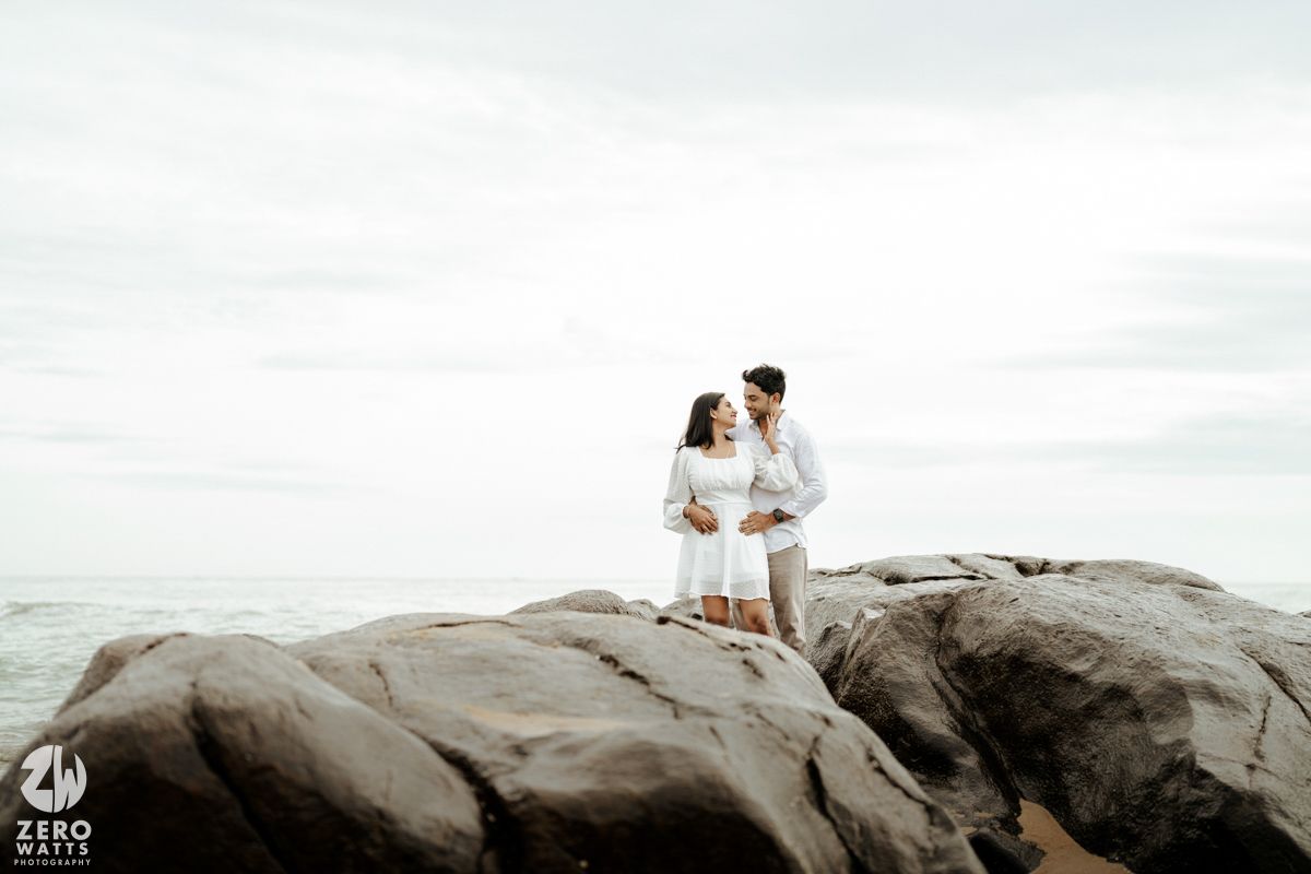 A couple in white outfits stands embracing on large coastal rocks against an overcast sky.