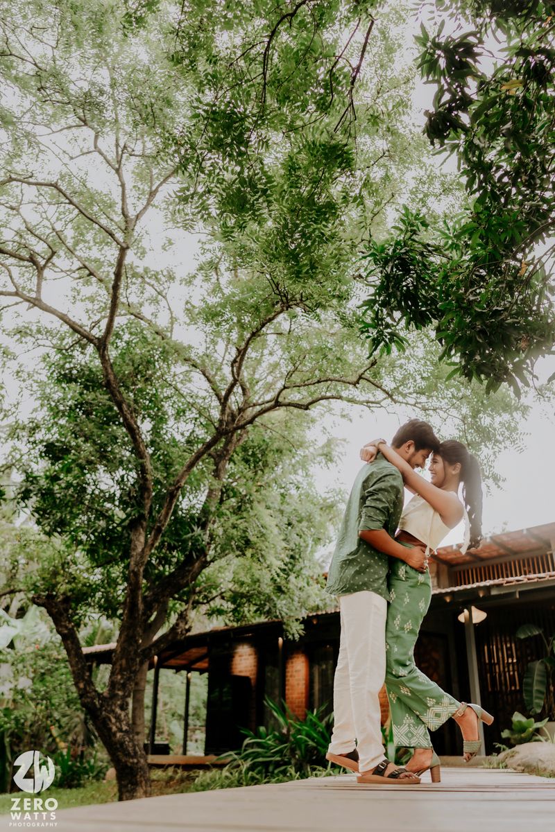 A couple enjoys a romantic moment beneath tall trees in a serene garden setting.