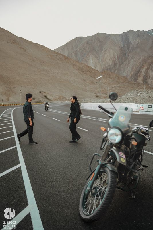 Two motorcyclists stand facing each other on a mountain road with their bike parked nearby.