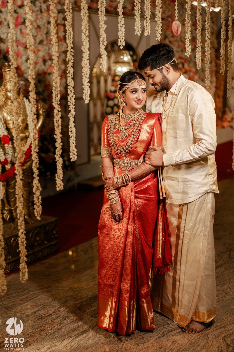 Traditional South Indian couple in wedding attire pose amid hanging floral decorations and lights.