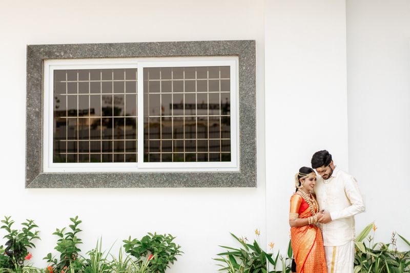 Couple in traditional Indian wedding attire posing in front of a white building with green plants.