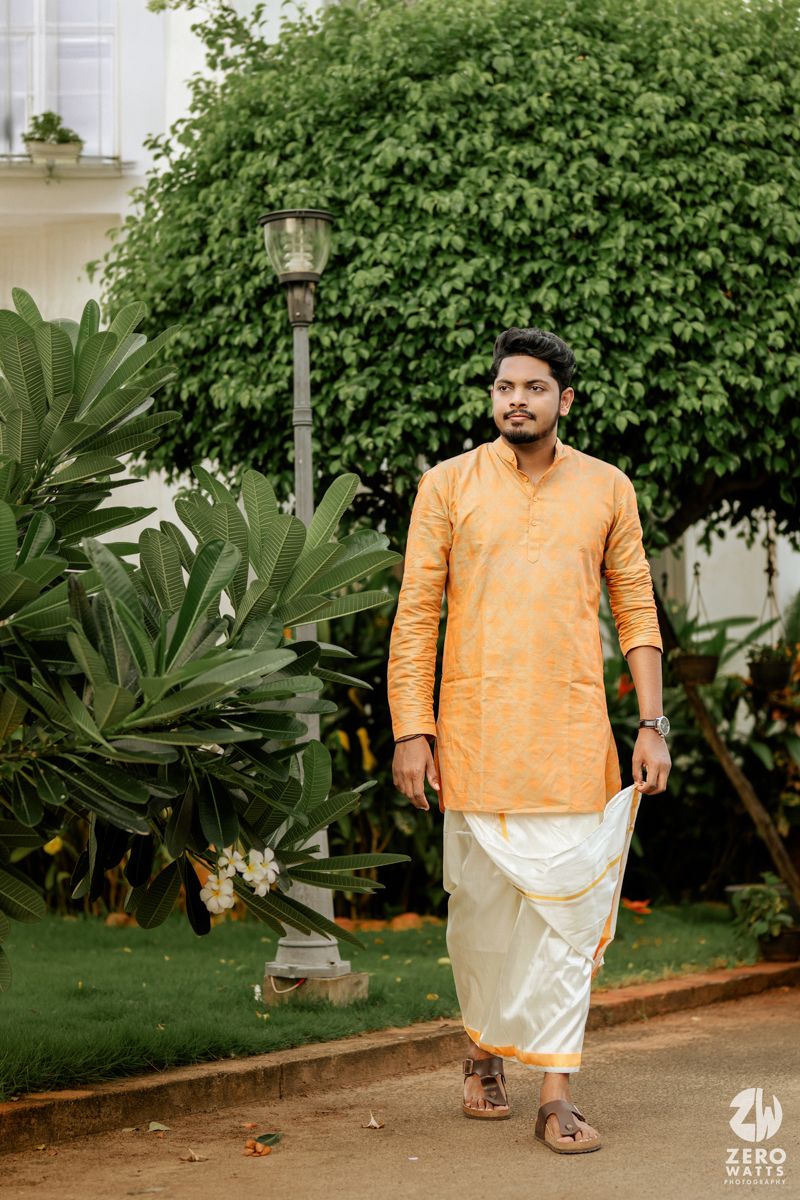 A person in traditional orange kurta and white dhoti stands on a garden path near lush greenery and a vintage lamp post.