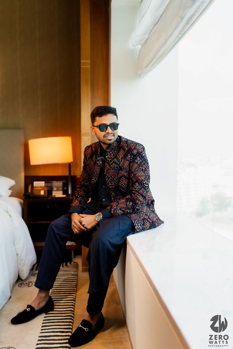Fashionable person in patterned jacket and sunglasses sits by hotel window with lamp and striped rug visible.
