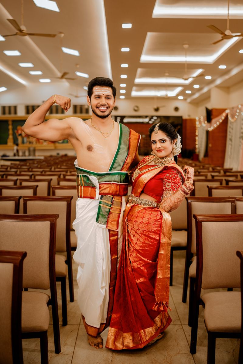 A couple poses in traditional South Indian wedding attire with the groom shirtless in a dhoti and bride in red silk saree.