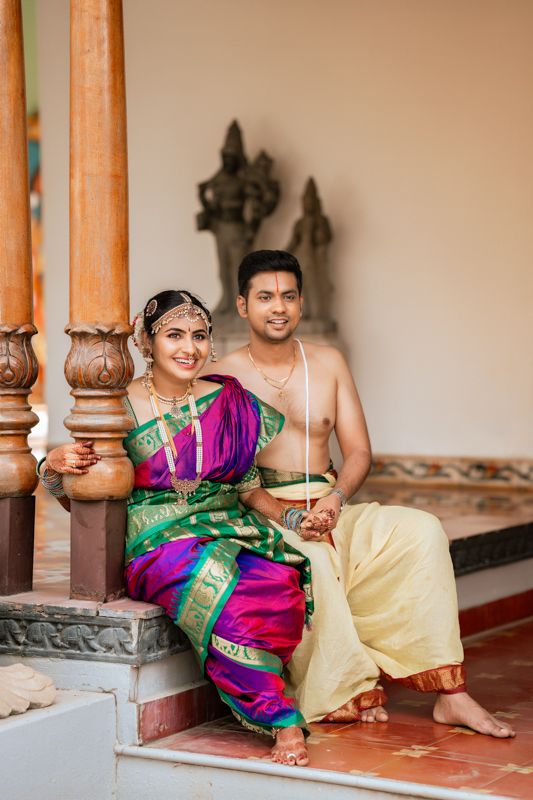 Couple in traditional South Indian wedding attire posing by wooden pillars in their ceremonial outfits.