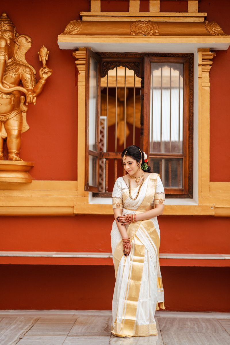 White and gold Kerala saree ensemble photographed against terracotta wall with traditional architectural details.