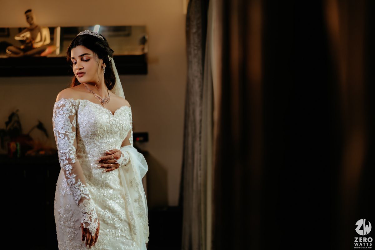 A bride in an elegant off-shoulder lace wedding dress with long sleeves stands in front of dark curtains.