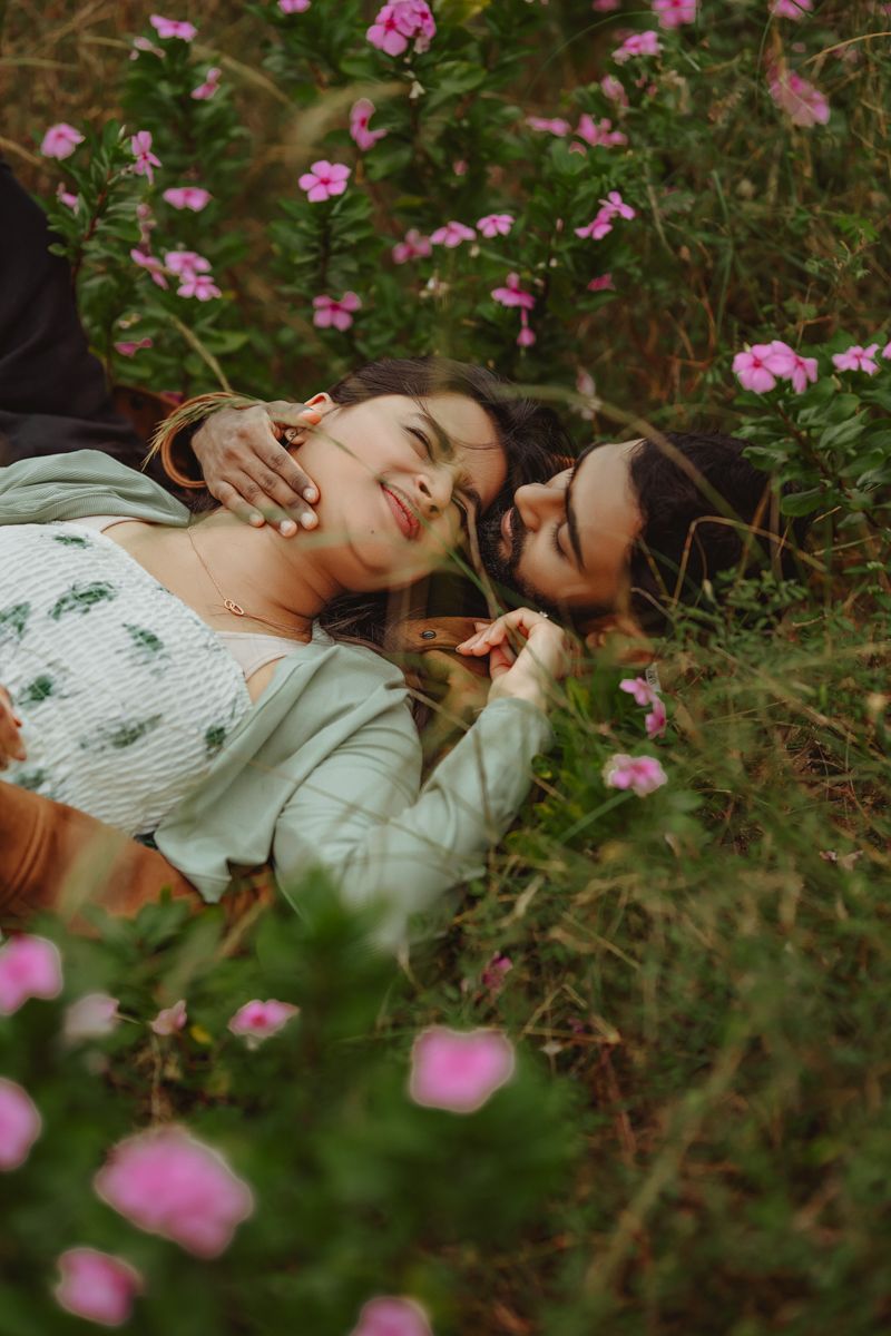 Two people lying among pink flowers and laughing together in a garden setting.