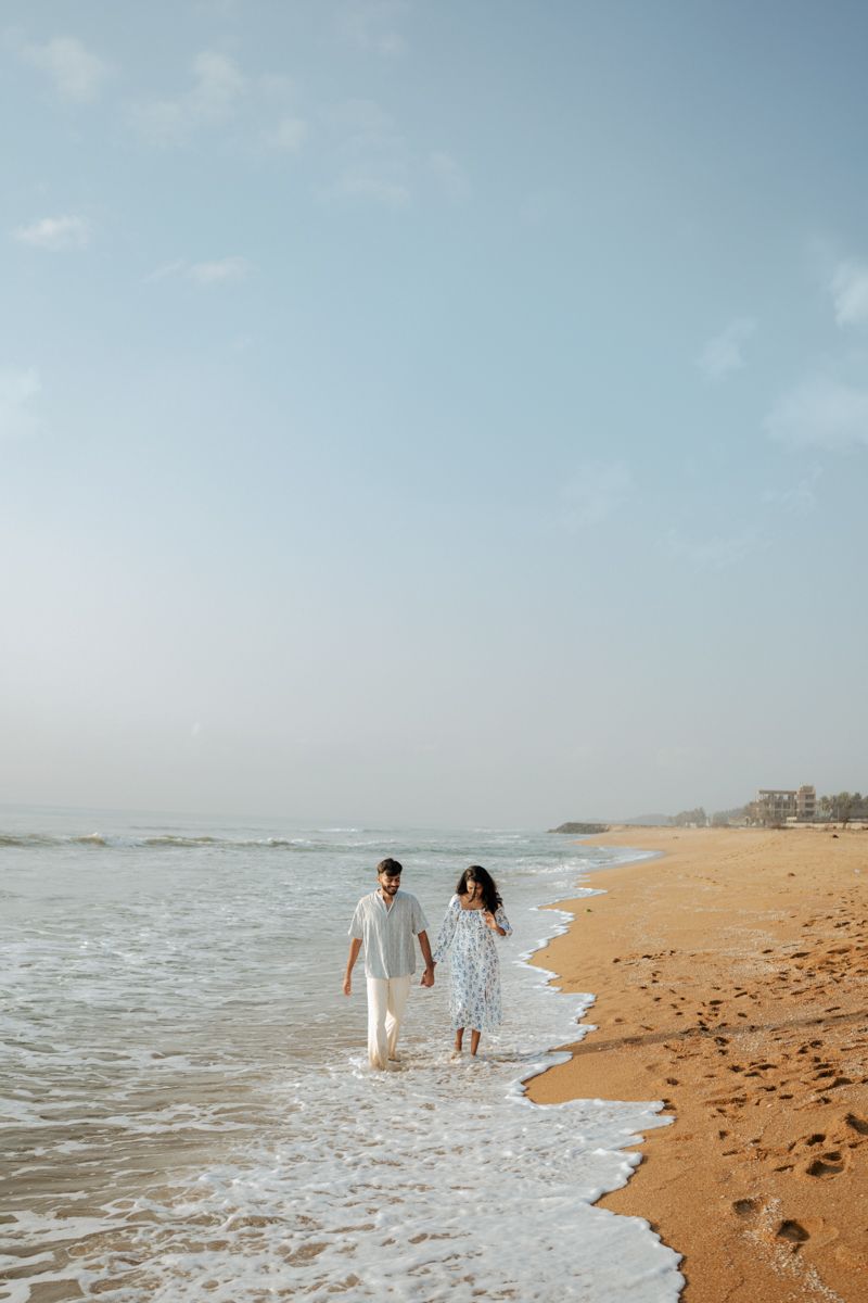 A couple walking hand in hand along a sandy beach with waves lapping at their feet.