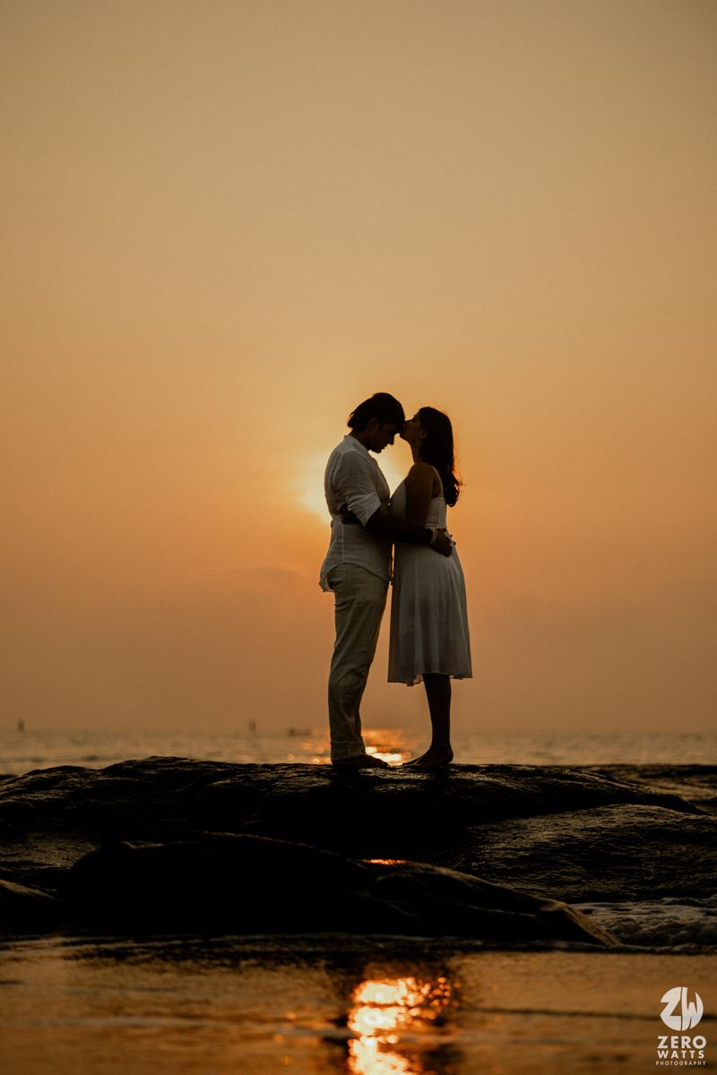 Silhouetted couple embraces on rocky beach during golden sunset with ocean waves in background.