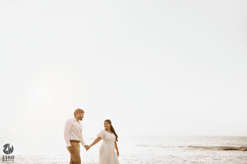 A couple holding hands while walking along the shoreline in elegant white and beige attire.