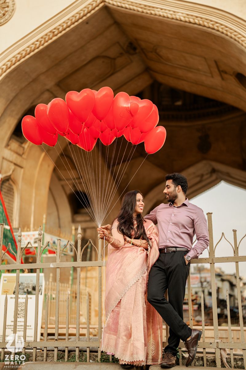 Couple poses with heart-shaped red balloons under ornate stone archway.