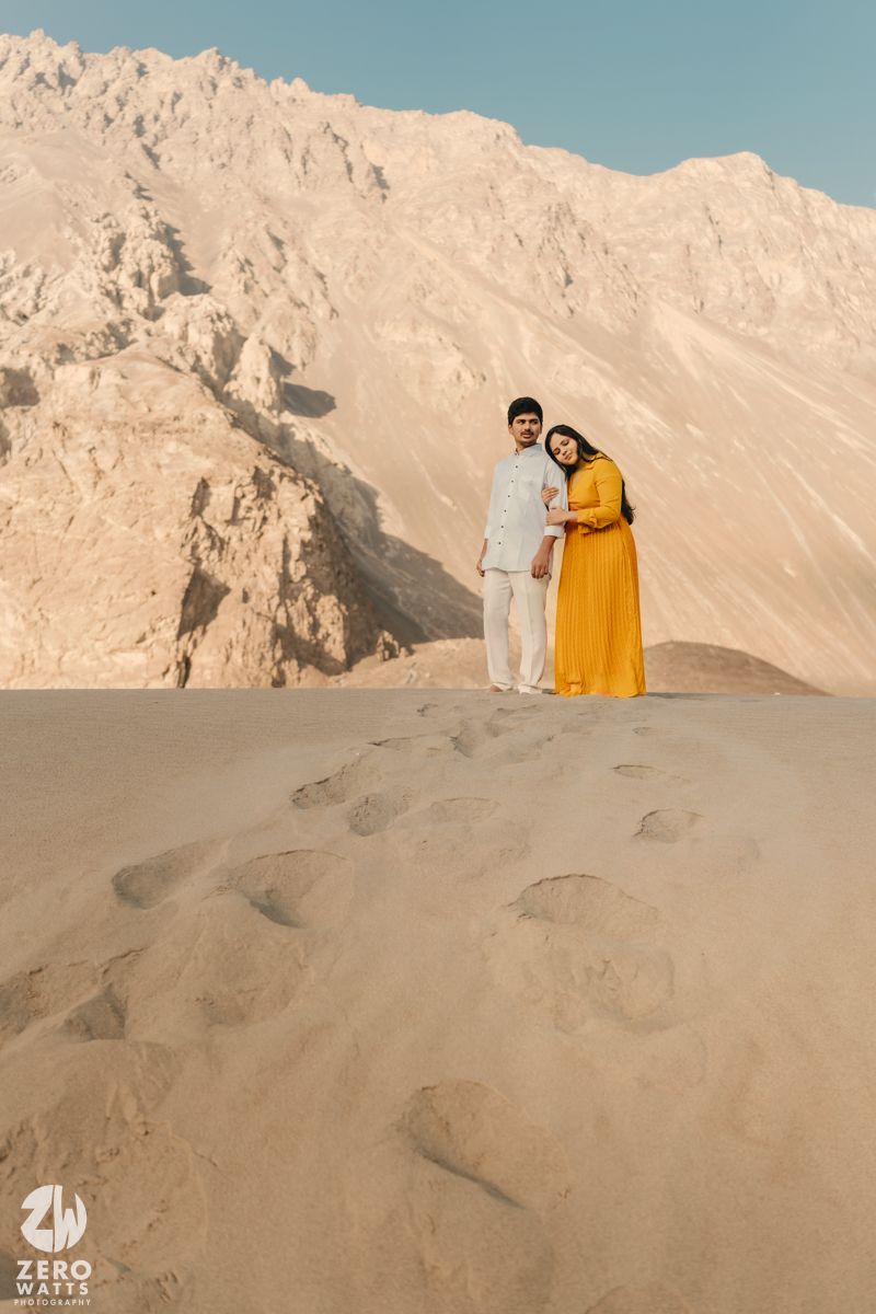 Two people in formal attire pose together on sandy dunes against rugged desert mountain landscape at golden hour.