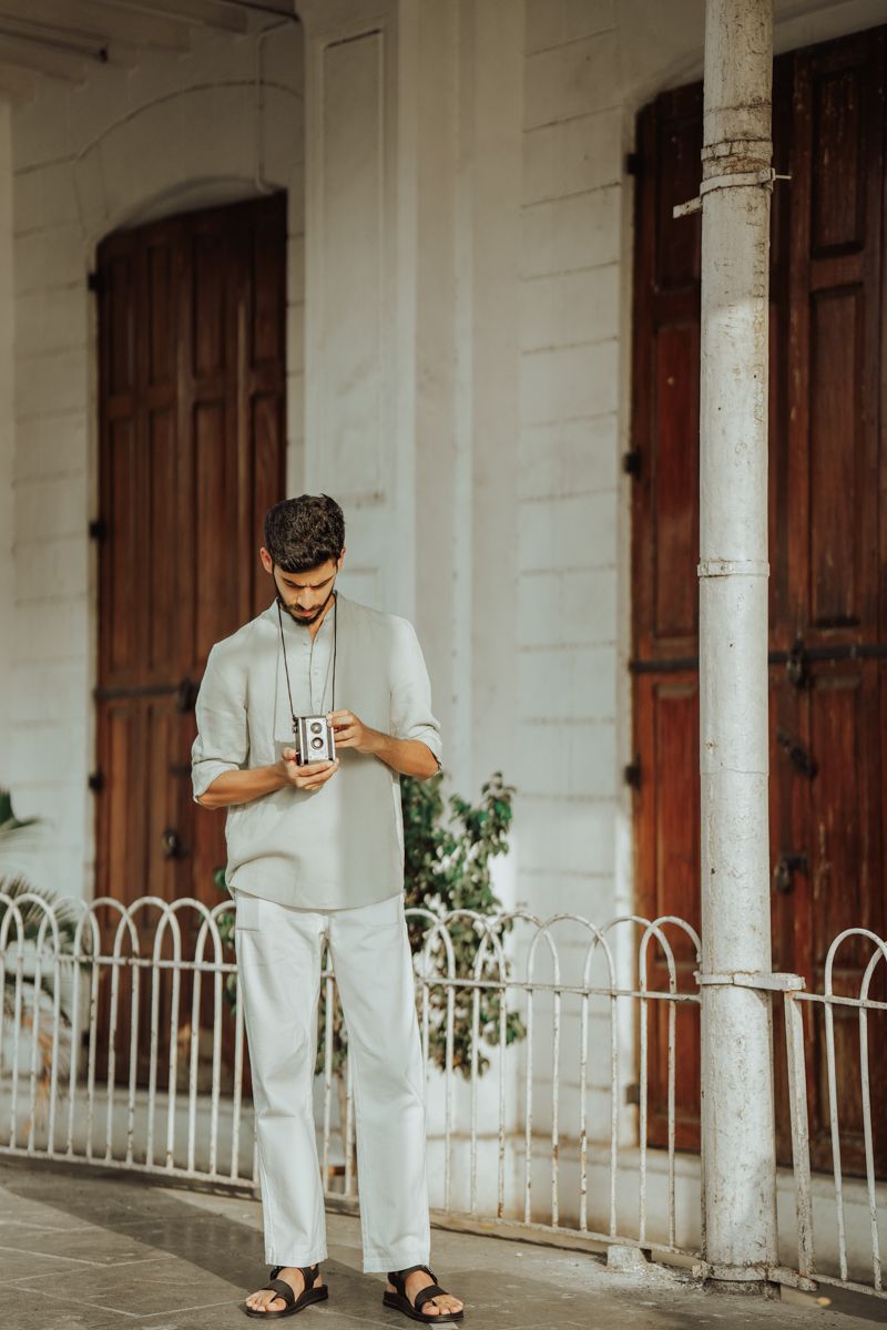 Person in casual white linen outfit checking phone while standing on a porch with wooden doors and white columns.