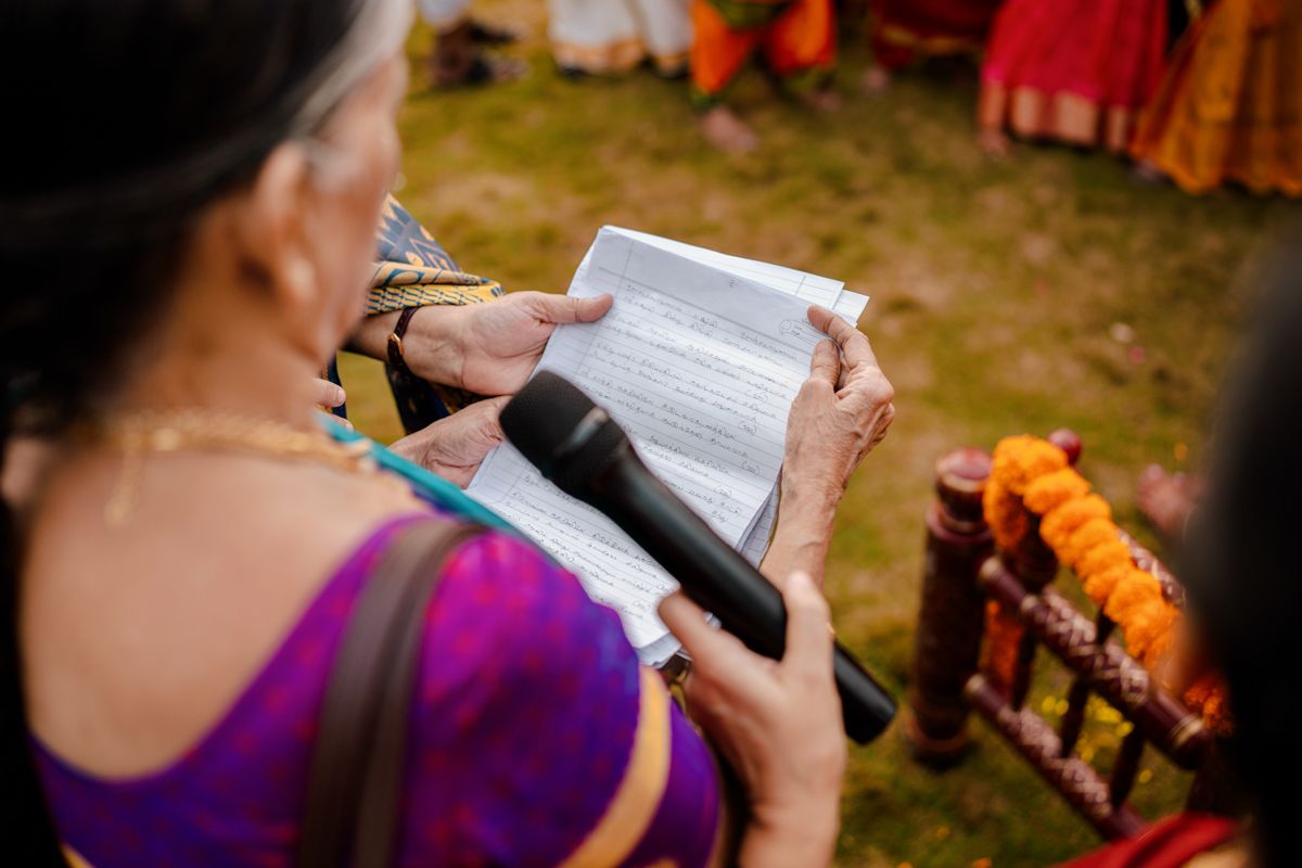 Someone in a purple traditional dress reads from a book during an outdoor cultural ceremony with decorative elements.