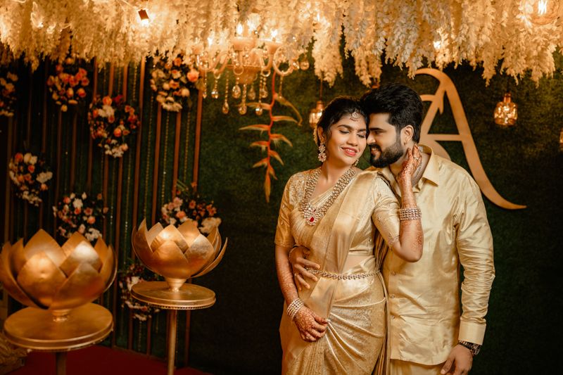 Romantic couple poses at elegant Indian wedding reception with gold decor, floral arrangements and hanging lights.