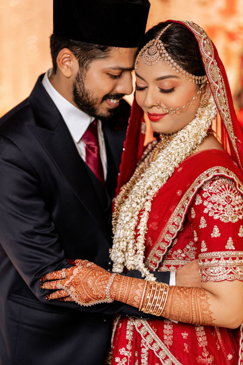 South Asian wedding couple in traditional attire with bride in red lehenga and groom in black suit sharing intimate moment.