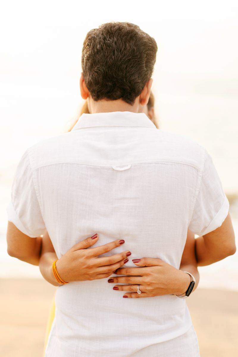 Close up view of hands with painted nails embracing someone in a white shirt from behind on a beach.