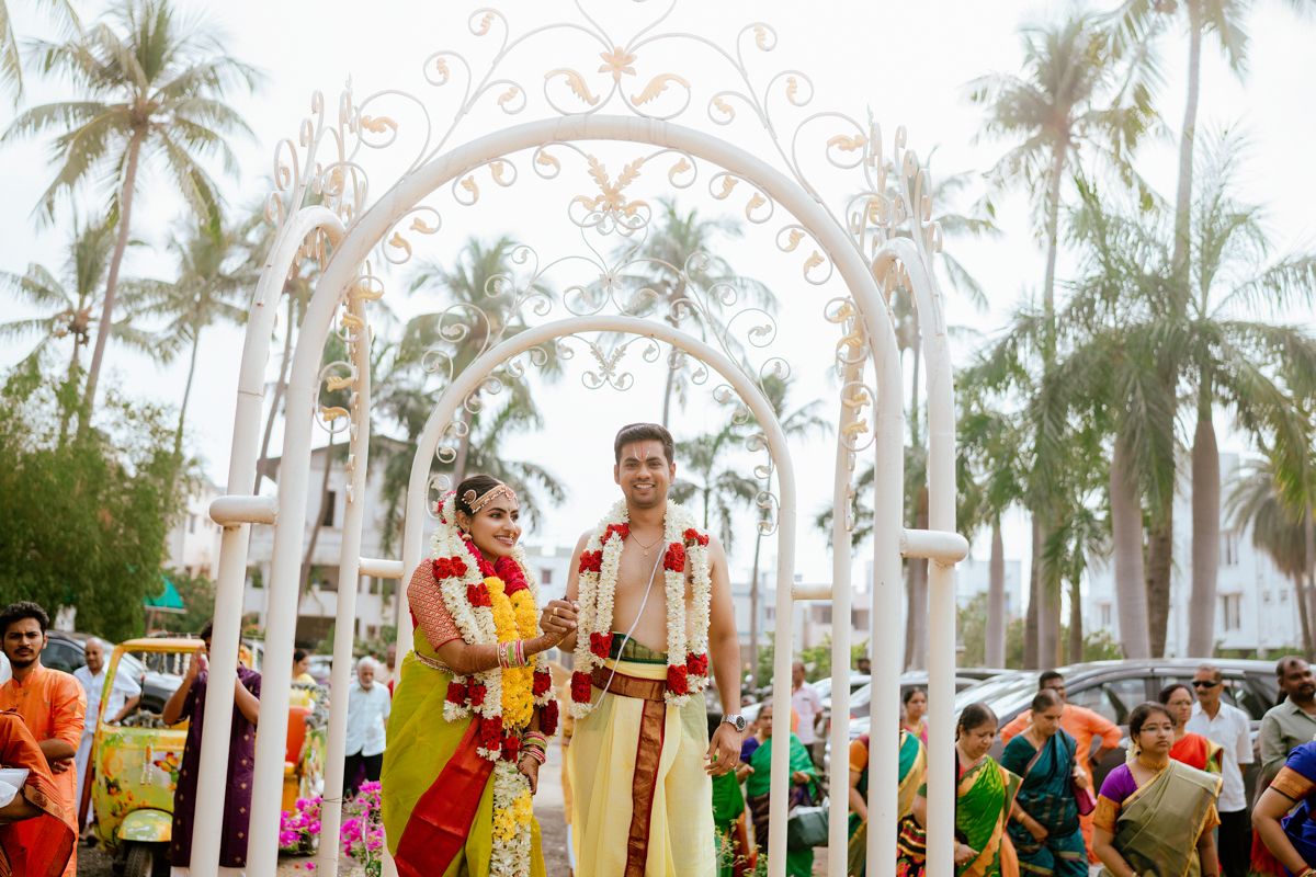 Traditional Indian wedding ceremony taking place under a white archway decorated with palm trees in the background.