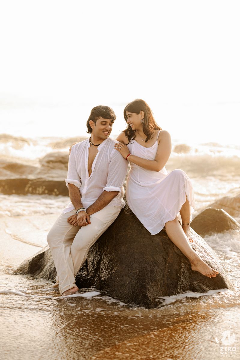 Couple in white outfits sitting together on a large rock at the beach during golden hour sunset.