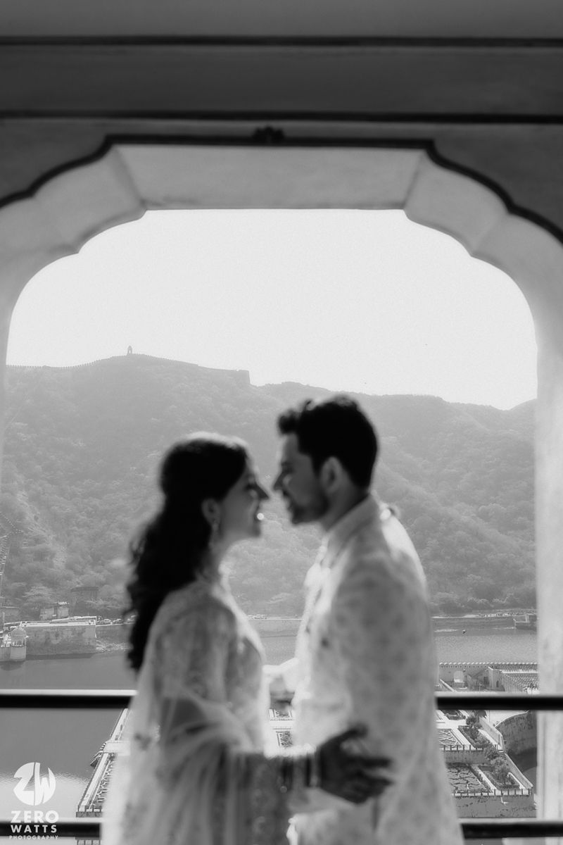 A black and white romantic scene of a couple in light clothing embracing by an arched window overlooking a scenic landscape.