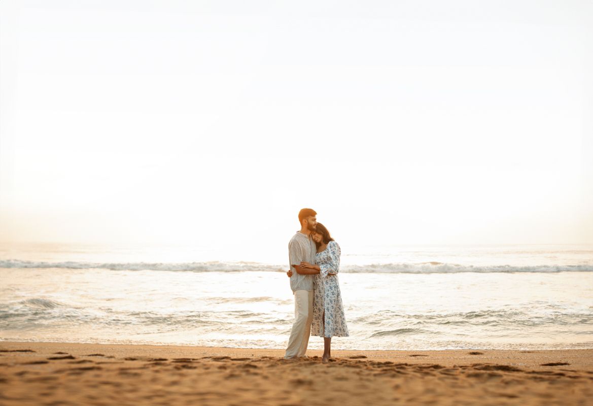 Couple in light colored clothing embracing on sandy beach at golden hour.