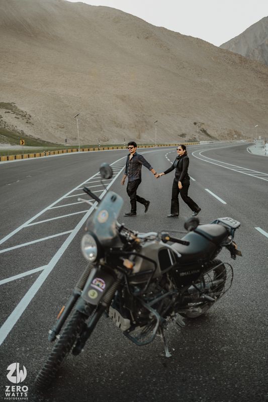 A motorcycle parked on a winding mountain road with two people holding hands against the desert landscape backdrop.