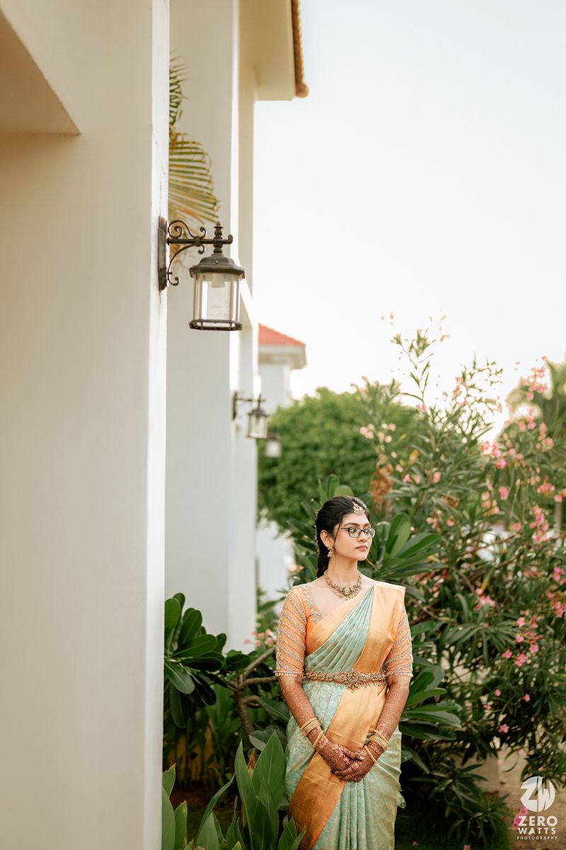 A person in a peach and mint green silk sari stands near tropical plants and an outdoor lantern at sunset.