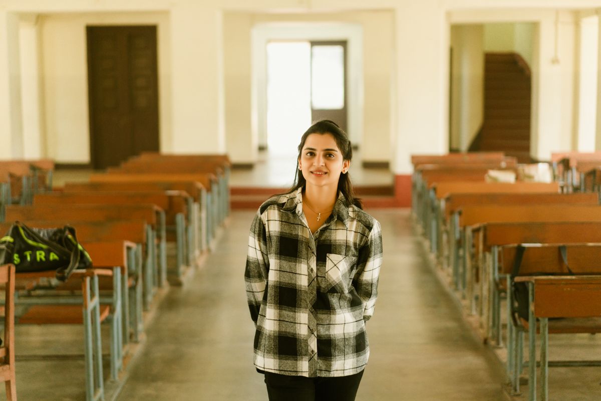Student standing in empty classroom with plaid flannel shirt and long dark hair smiling in the middle of a lecture hall.