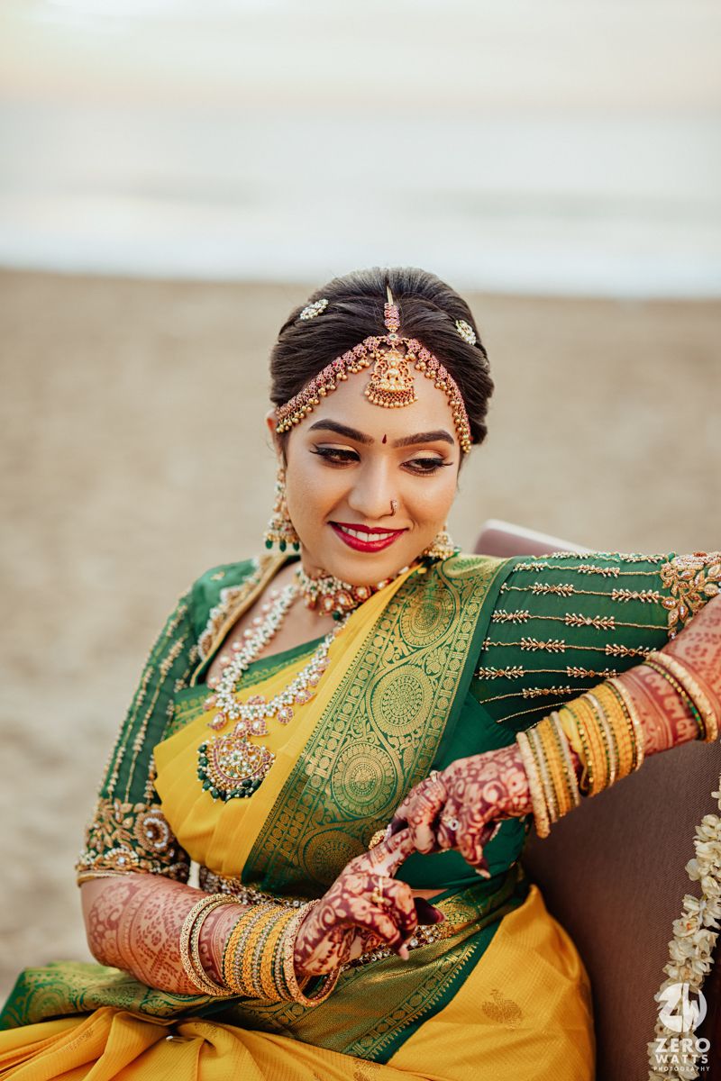 Traditional Indian bride wearing yellow and green silk saree with ornate jewelry sits on beach during wedding photoshoot.
