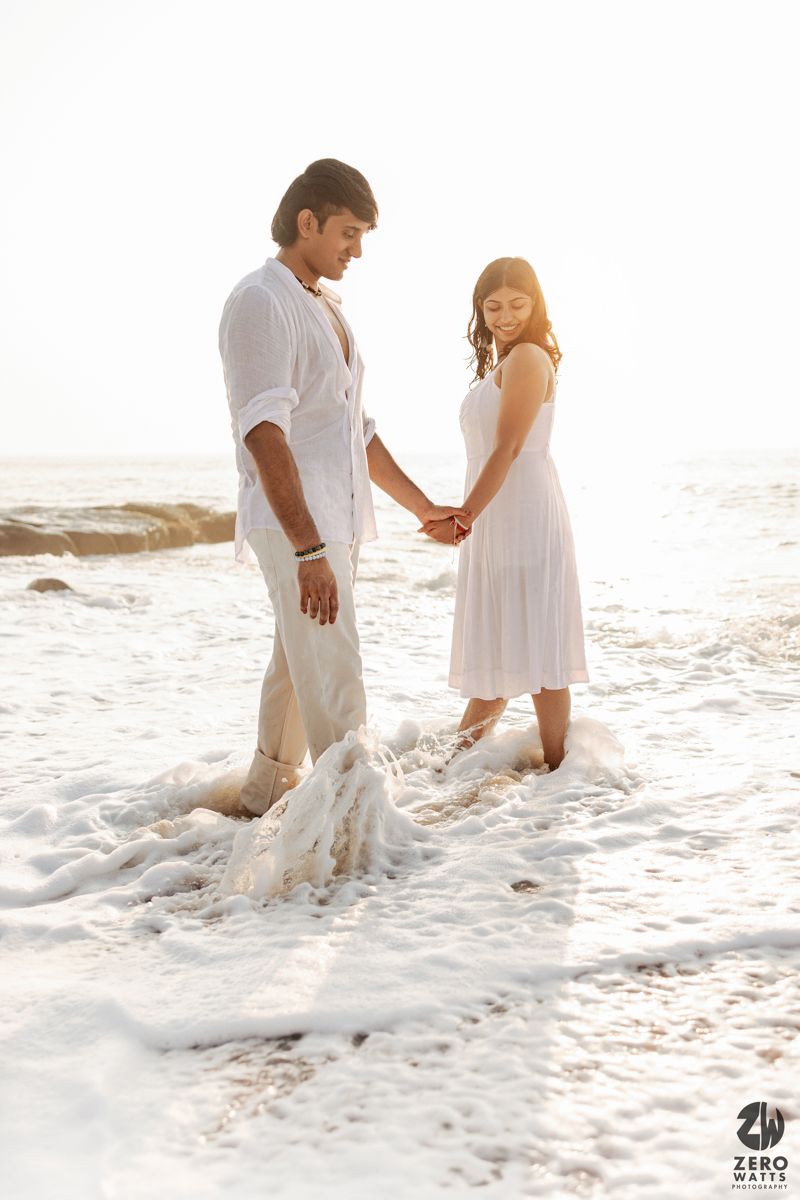 Couple in white outfits holding hands while standing in ocean waves at sunset on a romantic beach.