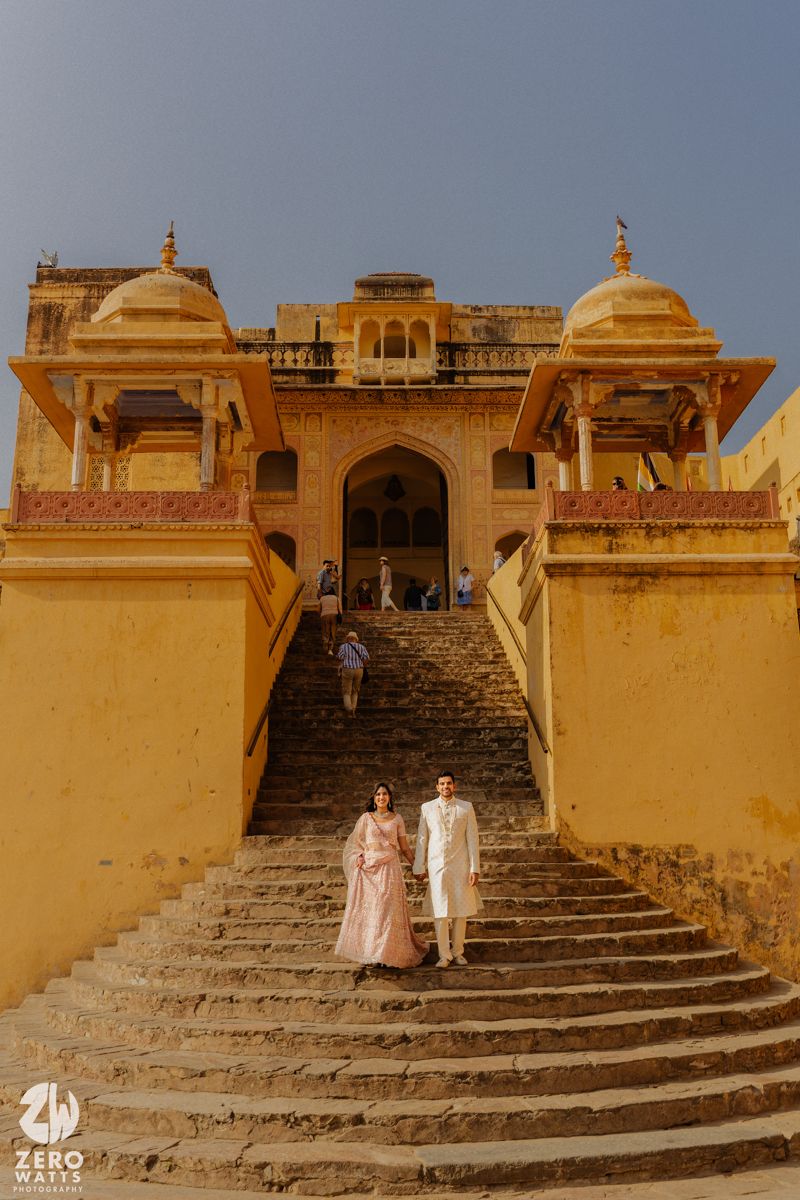 A couple in pink and white traditional Indian outfits stand on grand stone steps of a historic palace.