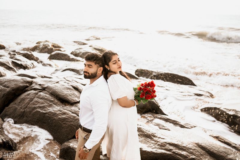 A couple poses against rocky coastal landscape holding red flowers during golden hour.