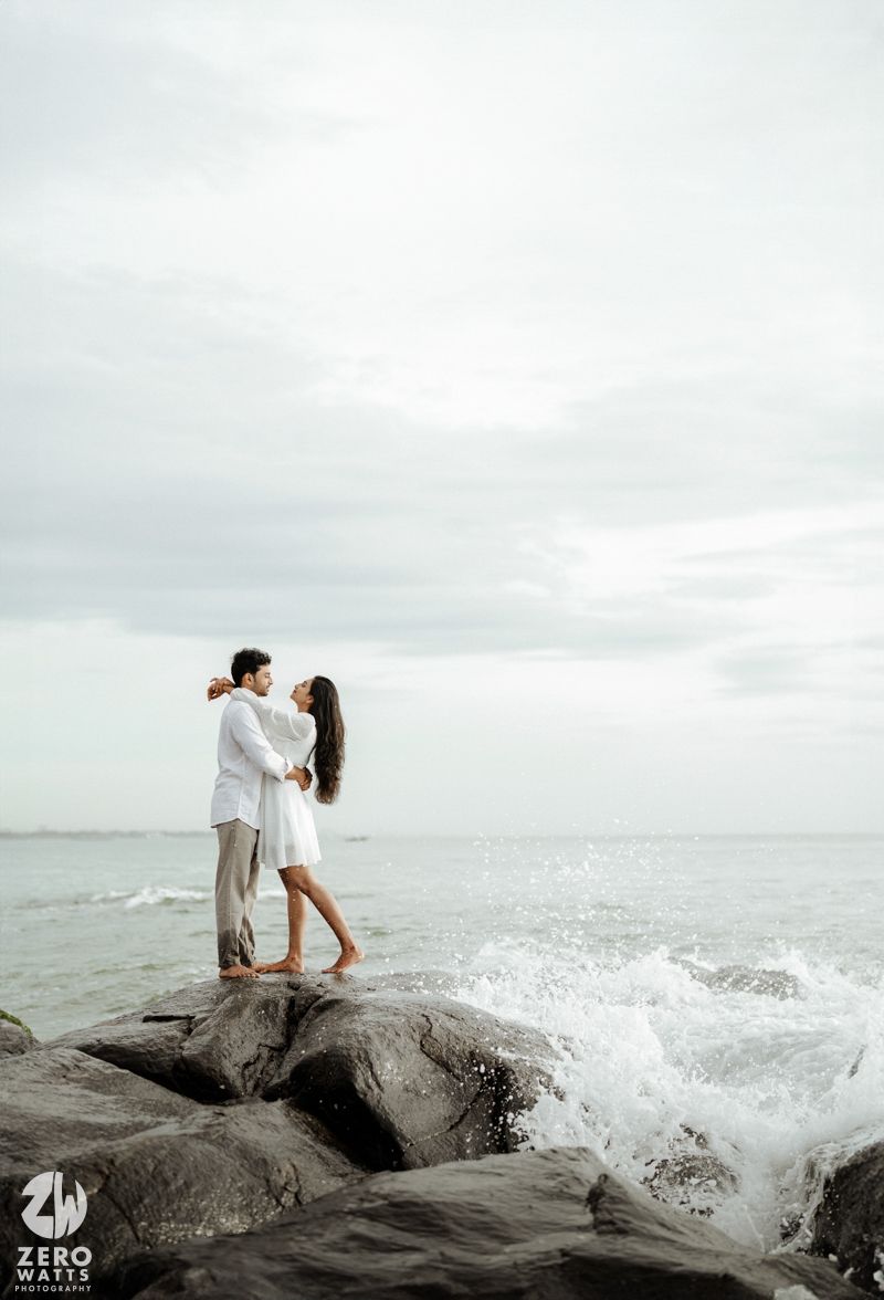 A romantic couple embraces on rocky coastal outcrop as ocean waves crash against the shore under an overcast sky.