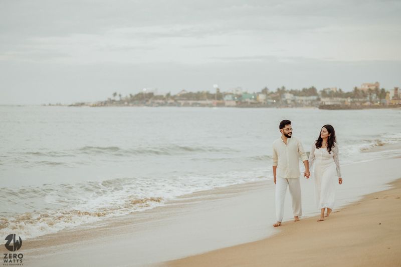 A romantic couple in white outfits taking a stroll along a serene beach during golden hour.