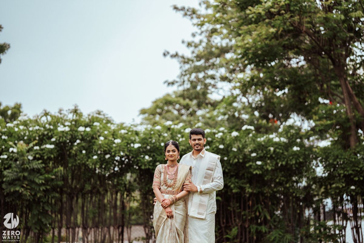 A couple stands together in traditional attire against a backdrop of white flowering bushes.