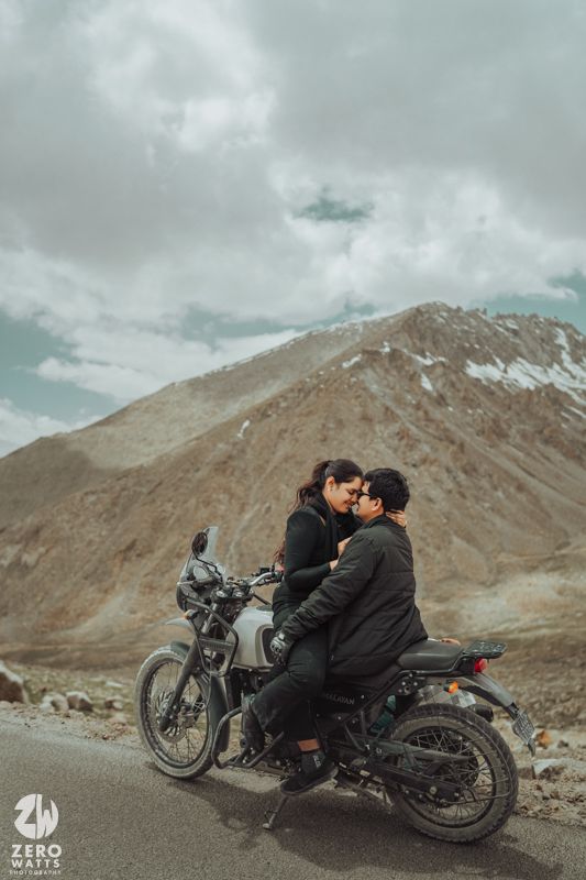 Two people share an intimate moment on a motorcycle against a backdrop of rugged mountain peaks and cloudy skies.