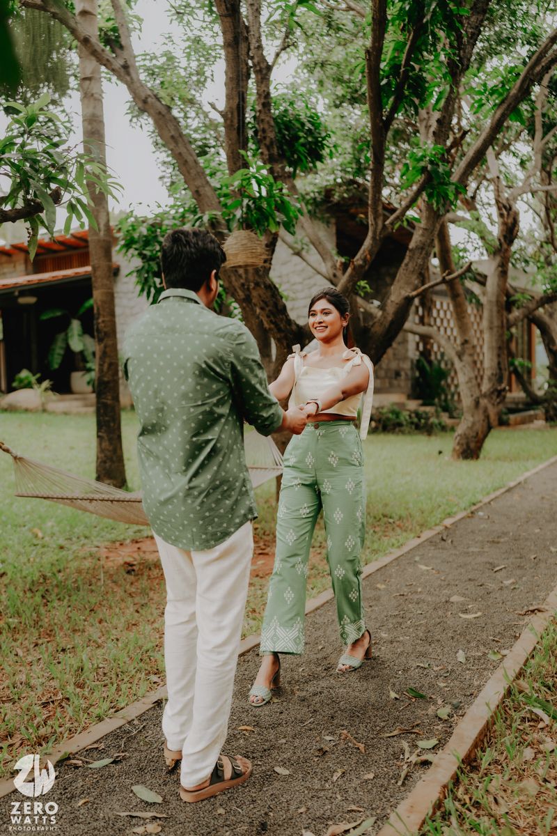 Two people standing beneath lush trees in a garden setting, wearing casual light-colored clothing.
