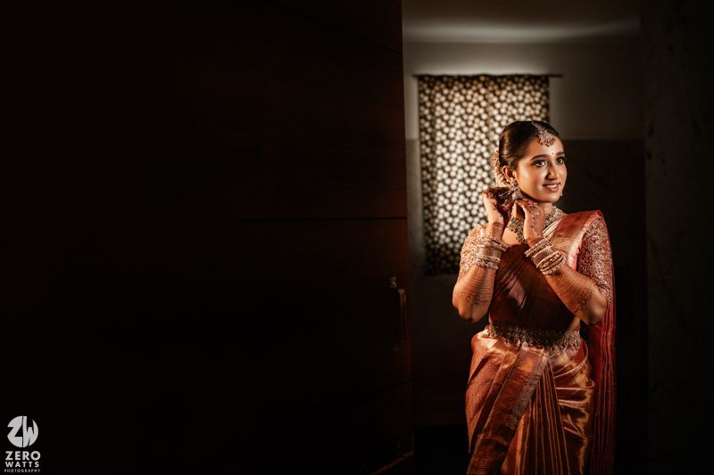 A person in a traditional silk saree stands in dramatic lighting against a dark room with a patterned curtain in the background.