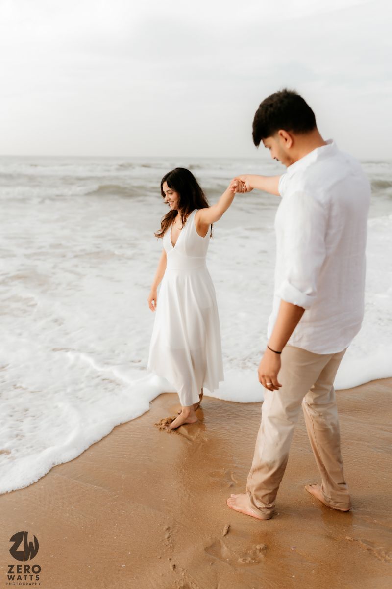 Couple playfully runs away from ocean waves on a sandy beach wearing white and beige outfits.